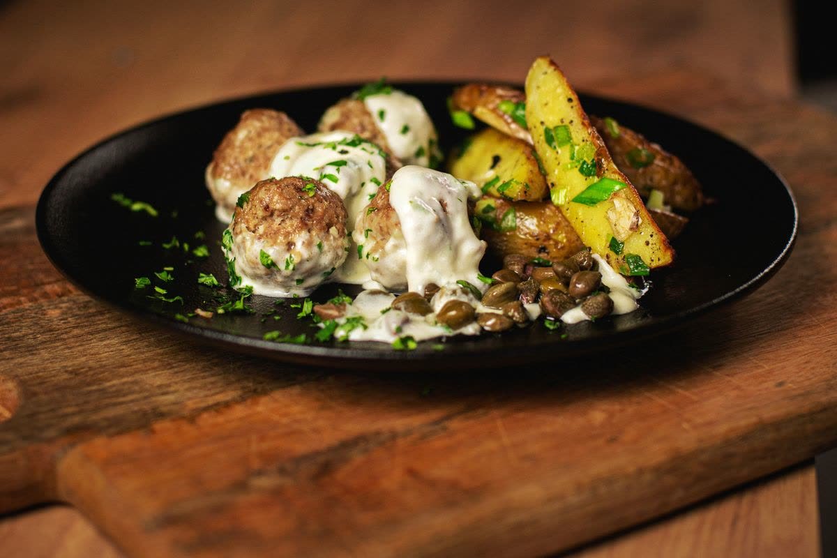 Köttbullar i krämig gräddsås serverade med klyftpotatis på en mörk tallrik.
