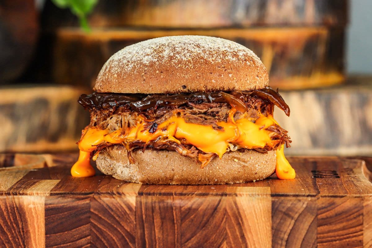 Macka med pulled pork och smält ost i ett grovt hamburgerbröd på en skärbräda.